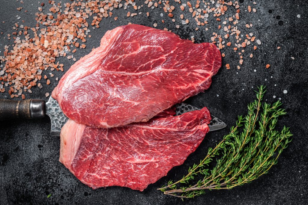 Butcher choise beef raw steaks on butcher knife. Black background. Top view